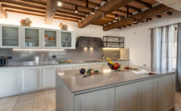 Modern kitchen interior featuring a spacious island with fresh vegetables, a stylish countertop, wooden beams, and elegant cabinetry. Natural light streams through the window, enhancing the inviting atmosphere.