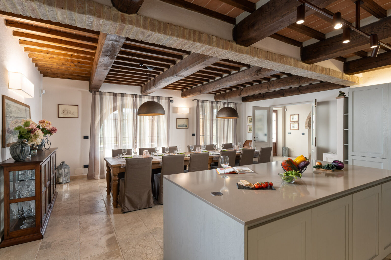 Spacious modern kitchen with wooden beams, featuring a large dining table, stylish pendant lights, and a central island adorned with fresh fruits. Natural light floods the room through large windows, creating a warm and inviting atmosphere. | Only on SopranoVillas