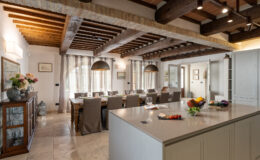 Spacious modern kitchen with wooden beams, featuring a large dining table, stylish pendant lights, and a central island adorned with fresh fruits. Natural light floods the room through large windows, creating a warm and inviting atmosphere.