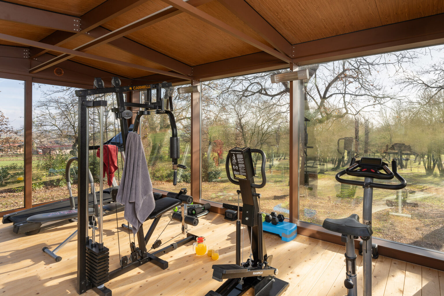Home gym interior featuring various fitness equipment including a weight machine, exercise bike, and free weights, with large windows offering a view of the outdoor landscape. | Only on SopranoVillas