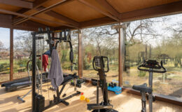 Home gym interior featuring various fitness equipment including a weight machine, exercise bike, and free weights, with large windows offering a view of the outdoor landscape.