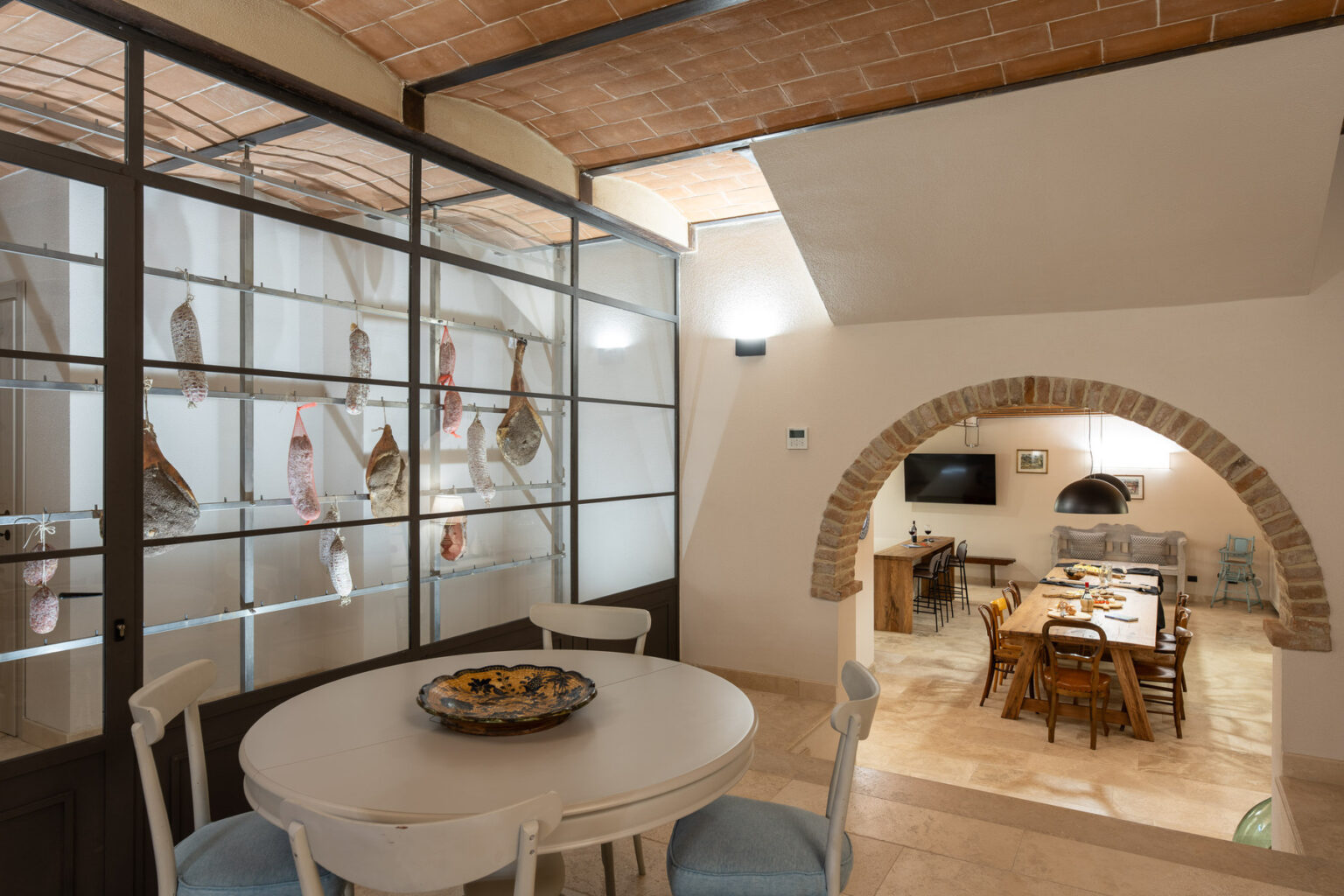 Interior view of a stylish dining area featuring a round table, decorated with a platter, and a glass wall showcasing cured meats. The space includes an arched entryway leading to a cozy kitchen with wooden dining furniture and a television. | Only on SopranoVillas