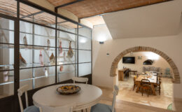 Interior view of a stylish dining area featuring a round table, decorated with a platter, and a glass wall showcasing cured meats. The space includes an arched entryway leading to a cozy kitchen with wooden dining furniture and a television.