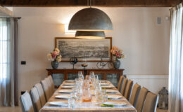 Elegant dining room featuring a long table set for a meal, with stylish tableware and floral centerpieces. A large pendant light hangs above the table, complemented by a decorative sideboard and artwork on the wall. Natural light filters in through sheer curtains, creating a warm and inviting atmosphere.