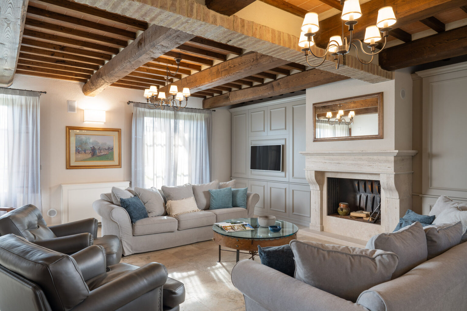 Modern living room with a cozy design featuring a beige sectional sofa, leather armchairs, a glass coffee table, and a fireplace. The space is illuminated by stylish chandeliers and has wooden beams on the ceiling, complemented by large windows with sheer curtains. | Only on SopranoVillas