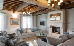 Modern living room with a cozy design featuring a beige sectional sofa, leather armchairs, a glass coffee table, and a fireplace. The space is illuminated by stylish chandeliers and has wooden beams on the ceiling, complemented by large windows with sheer curtains.