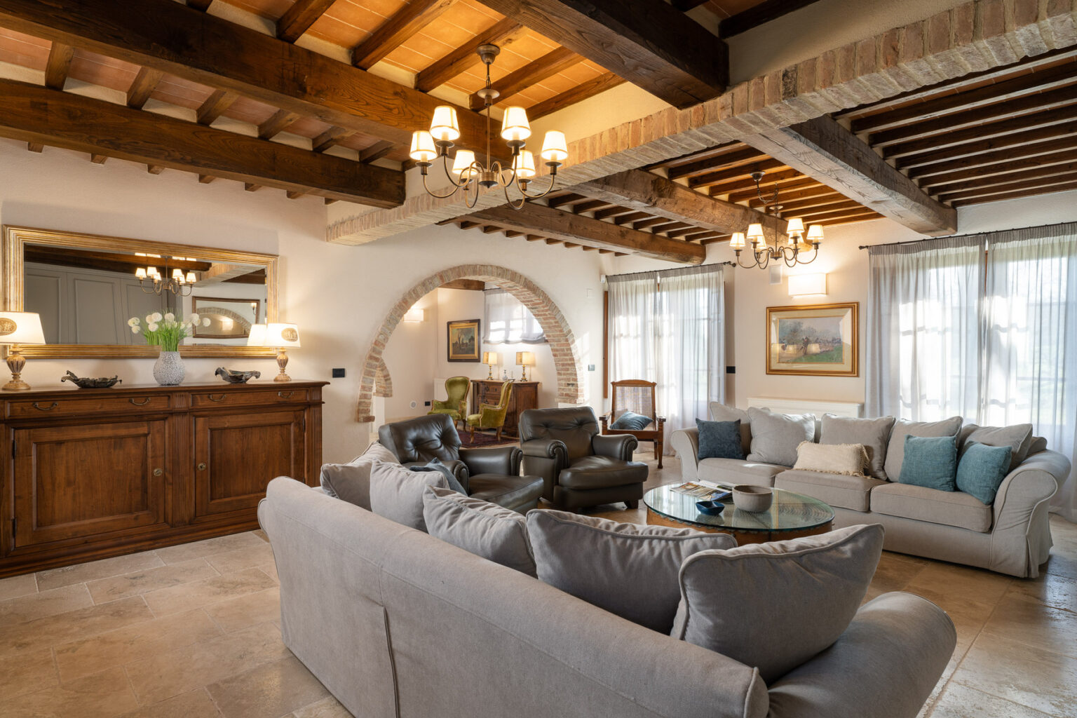 Spacious and elegantly designed living room featuring a mix of modern and rustic decor, with a comfortable gray sectional sofa, wooden beams on the ceiling, art pieces on the walls, and large windows allowing natural light to illuminate the space. | Only on SopranoVillas
