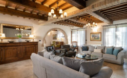 Spacious and elegantly designed living room featuring a mix of modern and rustic decor, with a comfortable gray sectional sofa, wooden beams on the ceiling, art pieces on the walls, and large windows allowing natural light to illuminate the space.
