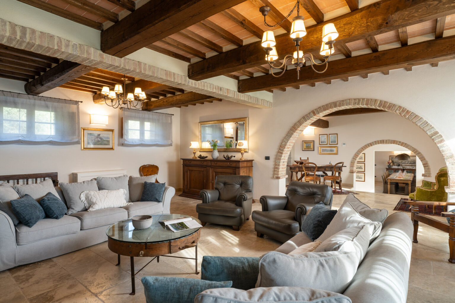 Spacious living room featuring a cozy arrangement of a light gray sofa and dark leather armchairs, complemented by a round glass coffee table. The room showcases rustic wooden beams, elegant chandeliers, and soft curtains, with an archway leading to a dining area adorned with framed artwork and a charming wooden sideboard. Natural light enhances the inviting atmosphere of this stylish interior space. | Only on SopranoVillas