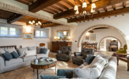 Spacious living room featuring a cozy arrangement of a light gray sofa and dark leather armchairs, complemented by a round glass coffee table. The room showcases rustic wooden beams, elegant chandeliers, and soft curtains, with an archway leading to a dining area adorned with framed artwork and a charming wooden sideboard. Natural light enhances the inviting atmosphere of this stylish interior space.