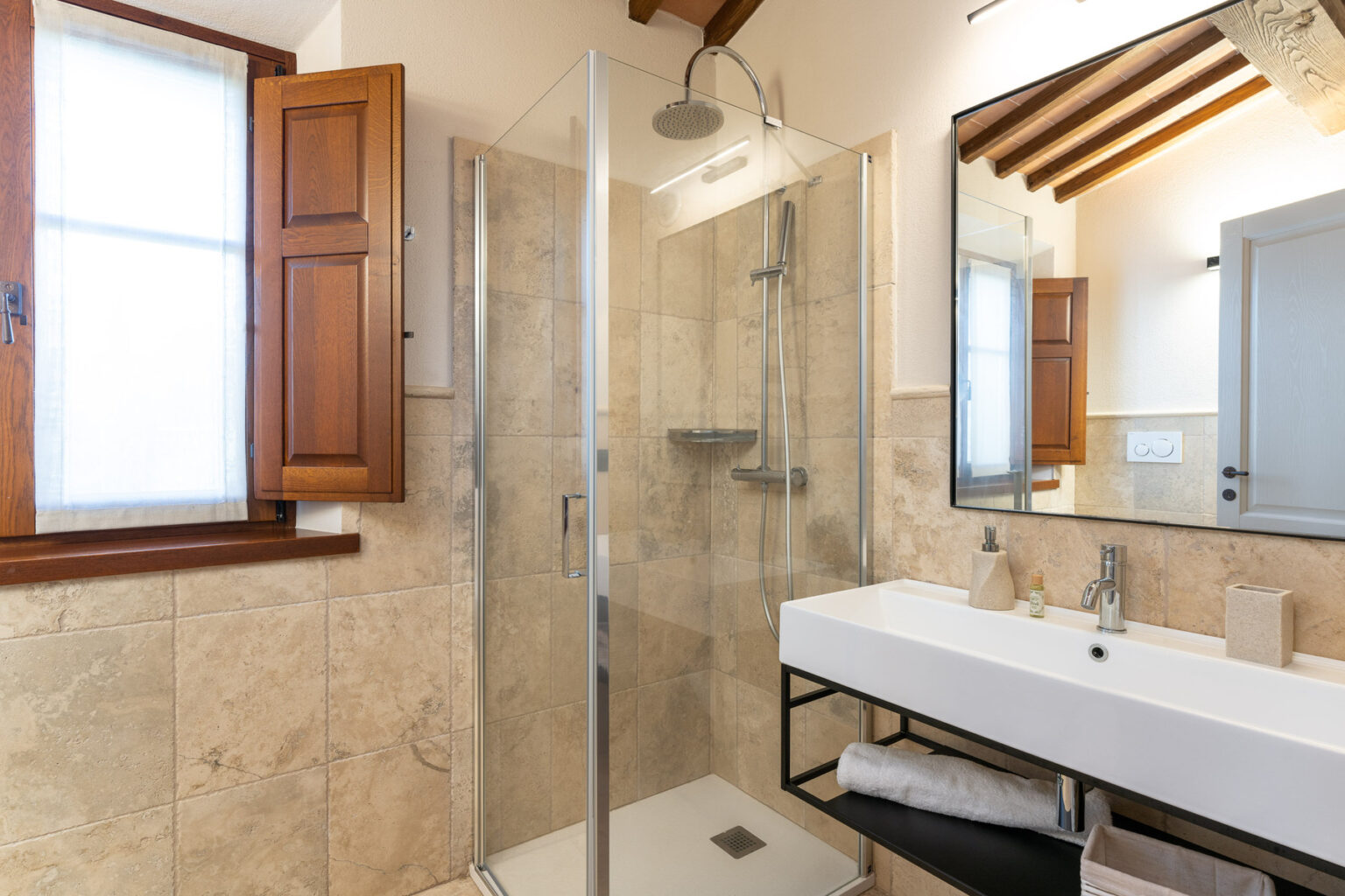 Modern bathroom featuring a glass shower enclosure, a sleek white sink with a black frame, and wooden window shutters. The design includes natural stone tiles, a large mirror, and stylish bathroom accessories, creating a sophisticated and inviting atmosphere. | Only on SopranoVillas