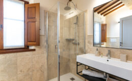 Modern bathroom featuring a glass shower enclosure, a sleek white sink with a black frame, and wooden window shutters. The design includes natural stone tiles, a large mirror, and stylish bathroom accessories, creating a sophisticated and inviting atmosphere.