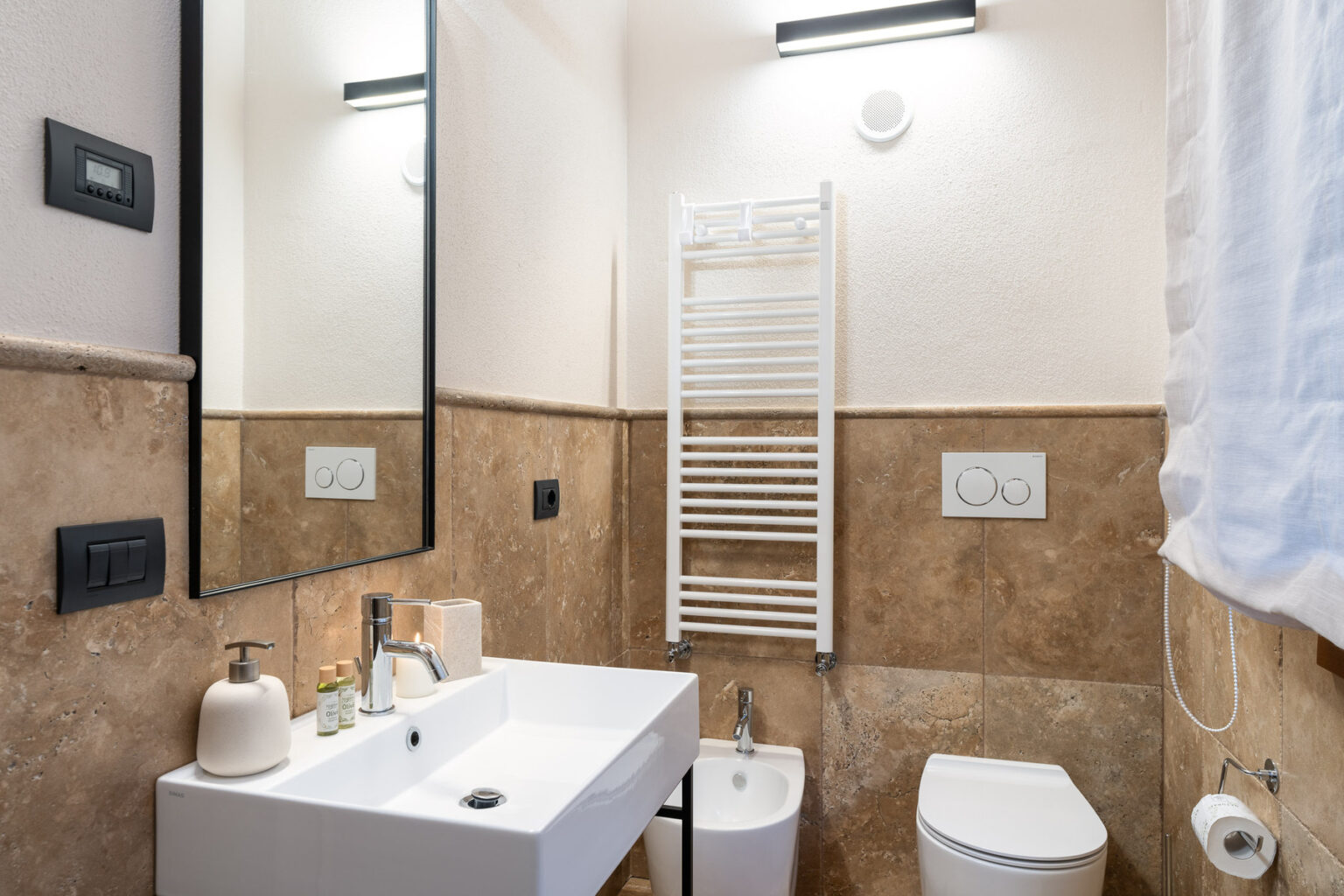 Modern bathroom featuring a sleek white sink with a contemporary faucet, a large mirror, and stylish stone walls. The room includes a towel radiator, a bidet, a toilet, and minimalist decor, creating an elegant and functional space. Soft lighting enhances the overall ambiance. | Only on SopranoVillas