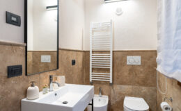 Modern bathroom featuring a sleek white sink with a contemporary faucet, a large mirror, and stylish stone walls. The room includes a towel radiator, a bidet, a toilet, and minimalist decor, creating an elegant and functional space. Soft lighting enhances the overall ambiance.