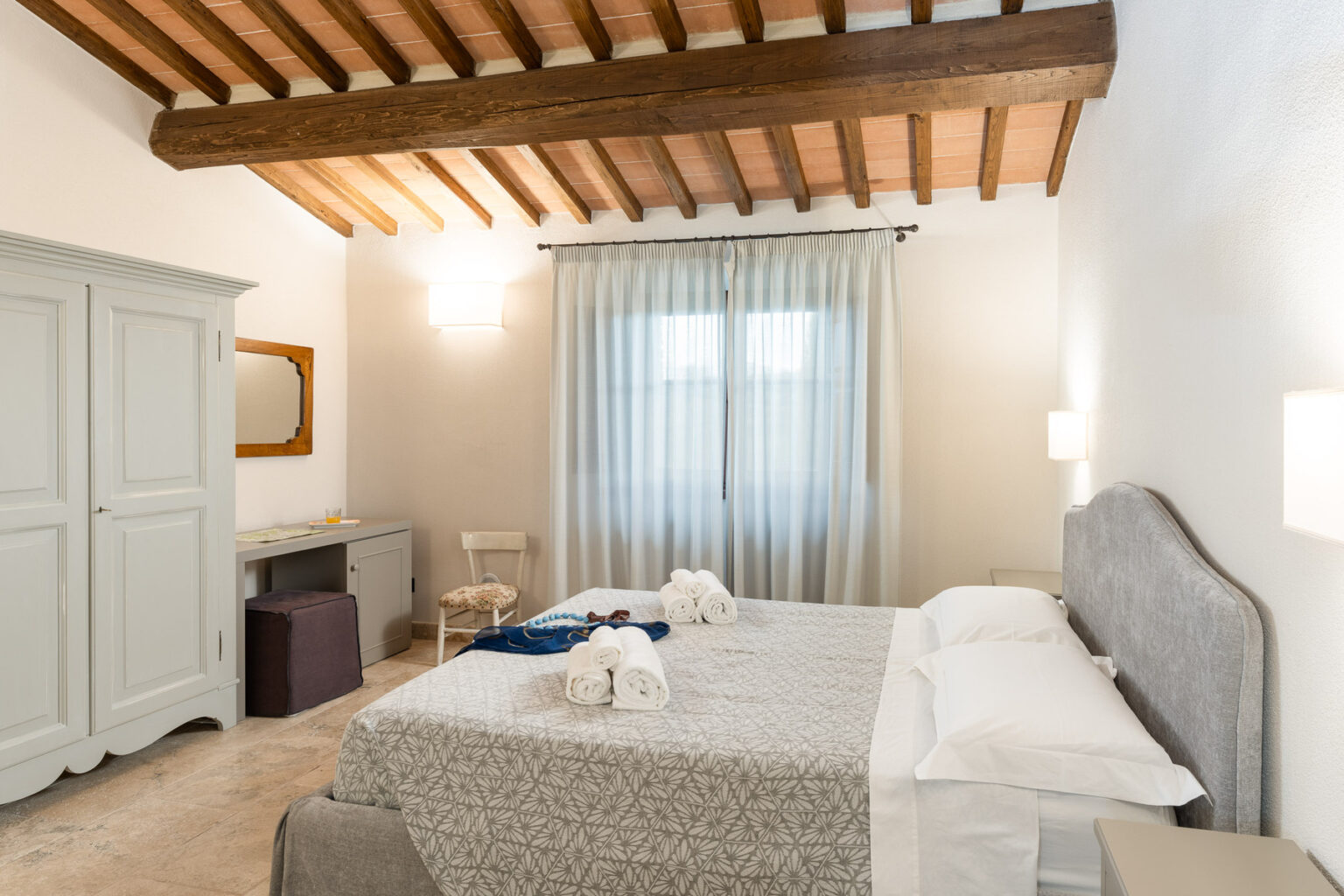 Cozy bedroom featuring a gray upholstered bed, elegant wooden ceiling beams, and soft natural light streaming through sheer curtains. The room includes a vintage-style wardrobe and a small dressing table, creating a warm and inviting atmosphere. | Only on SopranoVillas