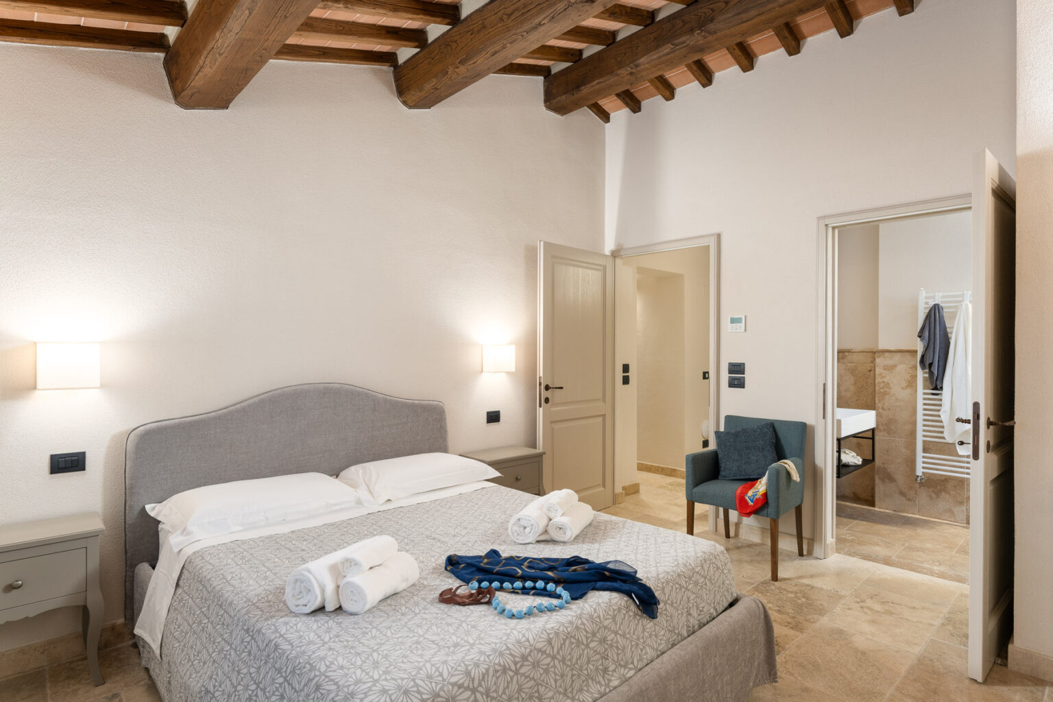 Cozy bedroom featuring a queen-sized bed with decorative pillows, two towels neatly arranged, a comfortable chair, and a warm color palette. The room showcases rustic wooden beams and modern decor, creating an inviting atmosphere. | Only on SopranoVillas