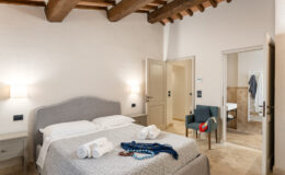 Cozy bedroom featuring a queen-sized bed with decorative pillows, two towels neatly arranged, a comfortable chair, and a warm color palette. The room showcases rustic wooden beams and modern decor, creating an inviting atmosphere.