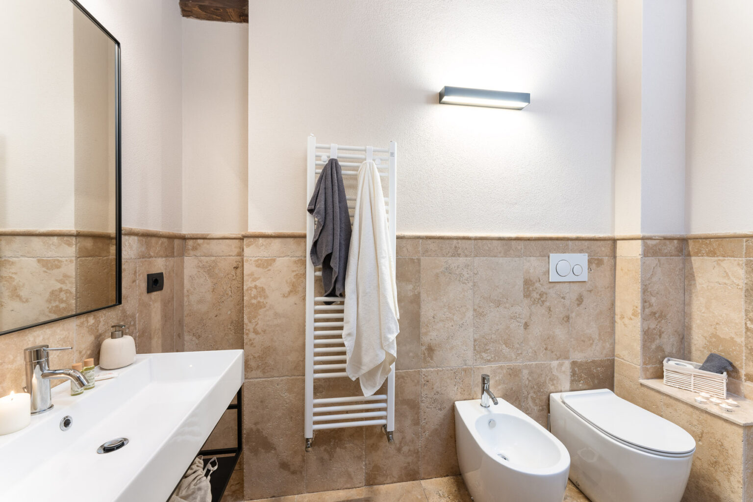 Modern bathroom interior featuring a sleek white sink, a bidet, and a towel rack with towels. The walls are adorned with light-colored tiles, and a large mirror reflects the ambient lighting. | Only on SopranoVillas
