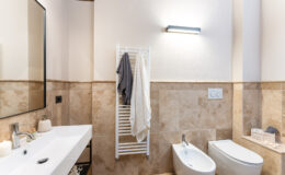 Modern bathroom interior featuring a sleek white sink, a bidet, and a towel rack with towels. The walls are adorned with light-colored tiles, and a large mirror reflects the ambient lighting.