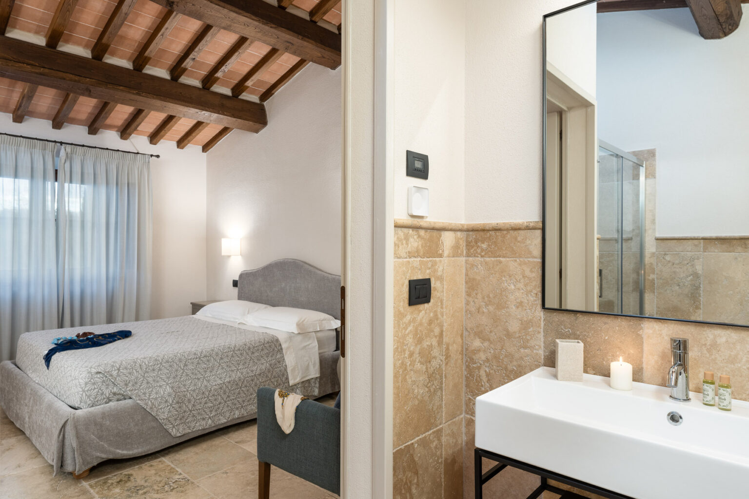 Cozy bedroom with wooden beams and large window, featuring a gray bedspread and soft lighting, adjacent to a modern bathroom with a sleek sink and glass shower. Perfect for a relaxing getaway. | Only on SopranoVillas