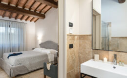 Cozy bedroom with wooden beams and large window, featuring a gray bedspread and soft lighting, adjacent to a modern bathroom with a sleek sink and glass shower. Perfect for a relaxing getaway.