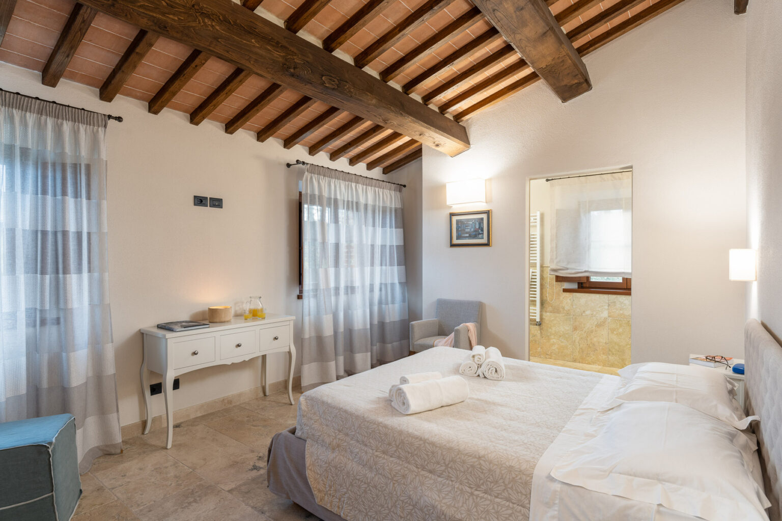 Cozy bedroom featuring wooden beams, soft curtains, a comfortable bed with neatly arranged linens, a stylish dresser, and an en-suite bathroom, creating a warm and inviting atmosphere. | Only on SopranoVillas