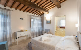 Cozy bedroom featuring wooden beams, soft curtains, a comfortable bed with neatly arranged linens, a stylish dresser, and an en-suite bathroom, creating a warm and inviting atmosphere.