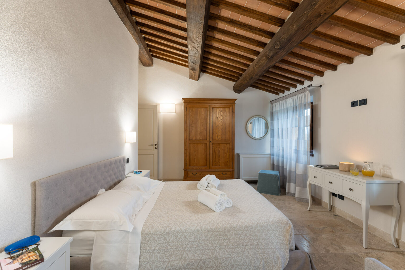 Cozy bedroom featuring a king-sized bed with a textured blanket, wooden beams on the ceiling, and natural light from large windows. The space includes a stylish dresser, a round mirror, and a wooden wardrobe, creating a welcoming and serene atmosphere. | Only on SopranoVillas