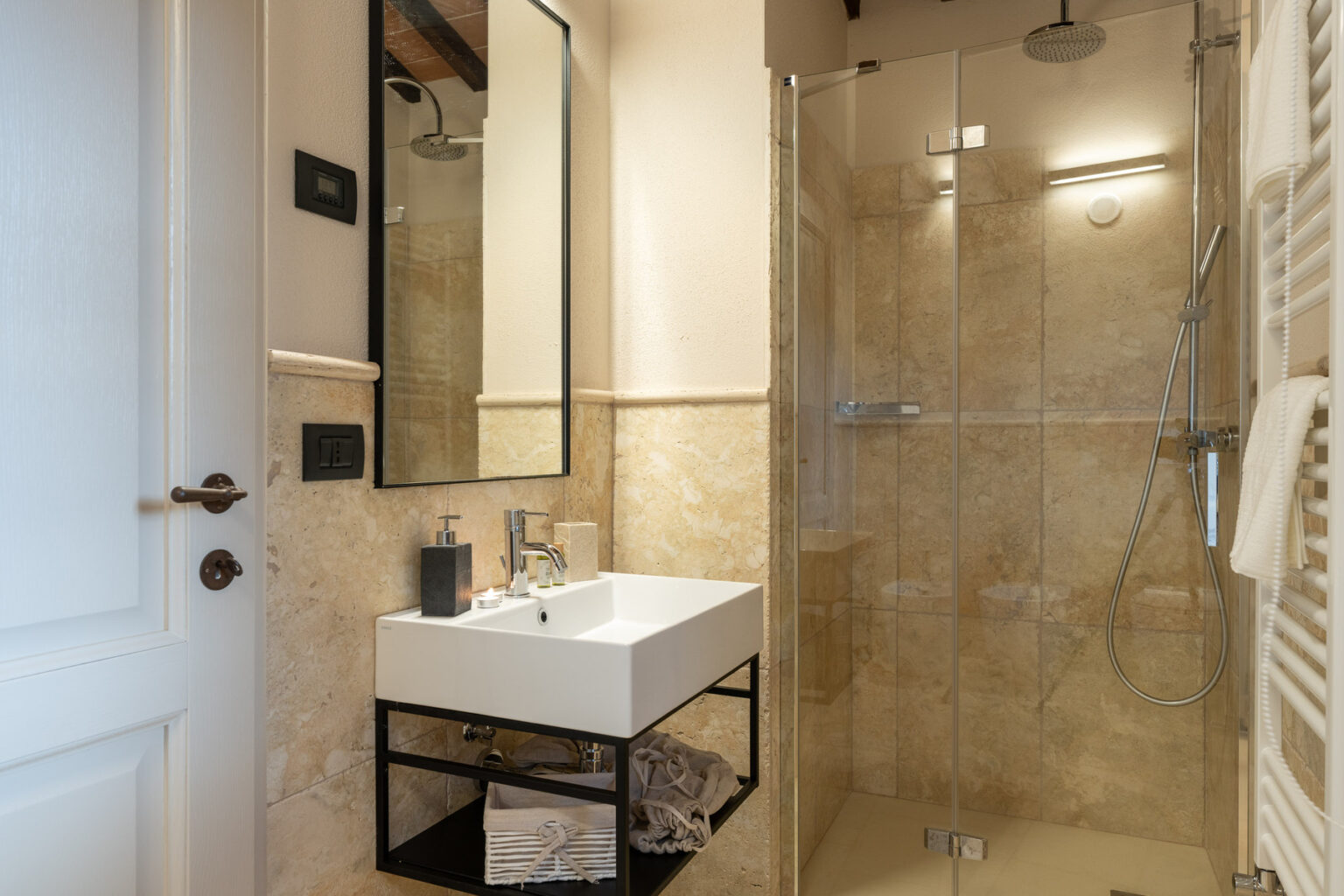 Modern bathroom featuring a glass shower enclosure, a sleek white sink on a black metal stand, and elegant beige stone walls. The space is well-lit and designed for functionality and style. | Only on SopranoVillas