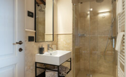 Modern bathroom featuring a glass shower enclosure, a sleek white sink on a black metal stand, and elegant beige stone walls. The space is well-lit and designed for functionality and style.