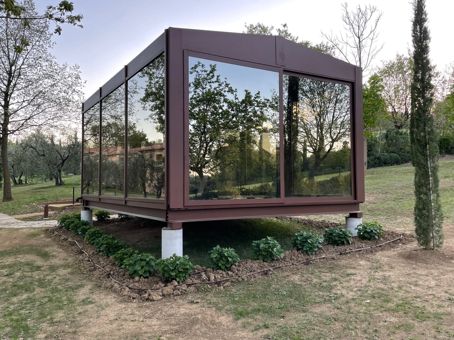 Modern glass house with a brown metal frame, situated in a landscaped garden with greenery. The structure features large reflective windows, showcasing surrounding trees and open space. Ideal for outdoor living and natural light. | Only on SopranoVillas