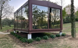 Modern glass house with a brown metal frame, situated in a landscaped garden with greenery. The structure features large reflective windows, showcasing surrounding trees and open space. Ideal for outdoor living and natural light.