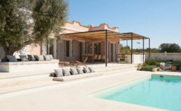 Modern villa with a swimming pool, featuring a spacious outdoor terrace and comfortable seating, surrounded by greenery under a clear blue sky.