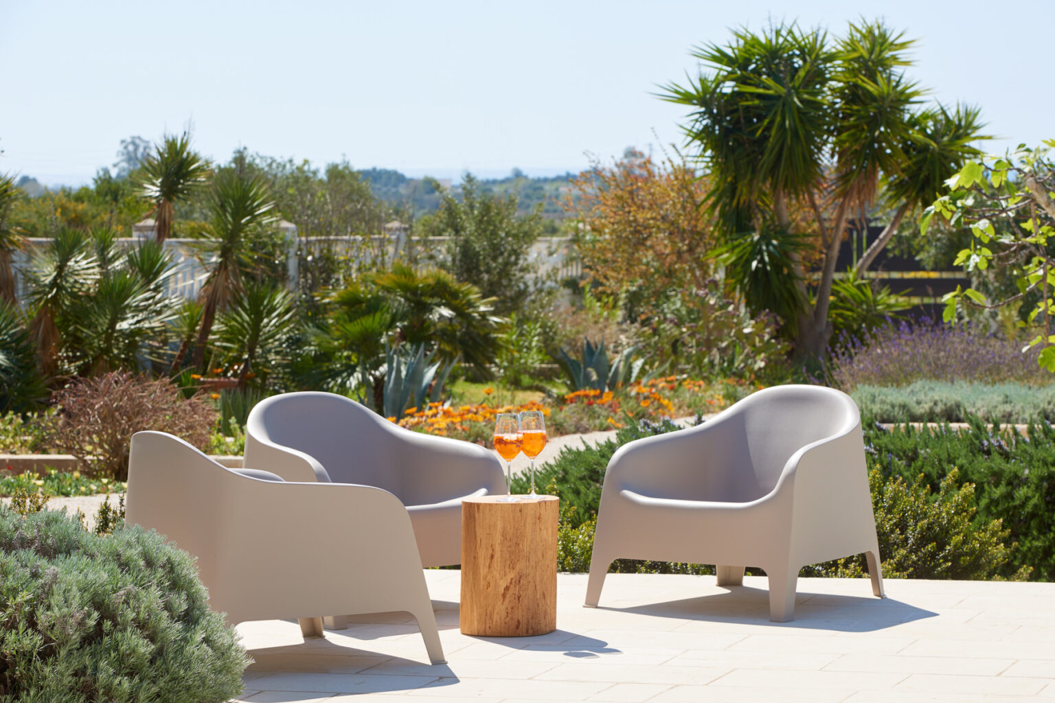 Two modern outdoor chairs in a serene garden setting, surrounded by lush greenery and colorful plants, with a wooden stump table in between. | Only on SopranoVillas