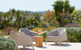 Two modern outdoor chairs in a serene garden setting, surrounded by lush greenery and colorful plants, with a wooden stump table in between.