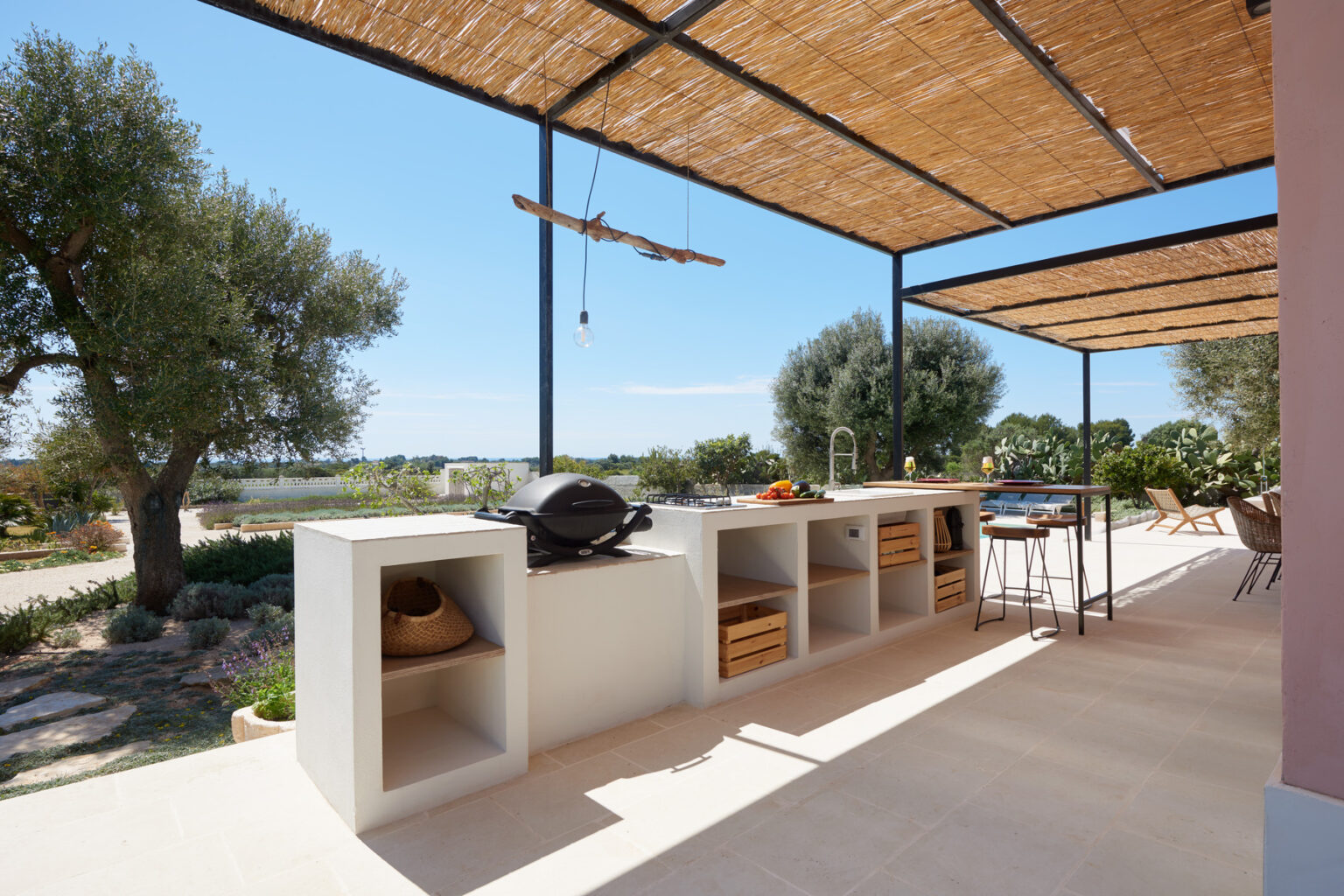 Outdoor kitchen area featuring a modern design with a stone grill and open storage spaces, set under a shaded pergola. Lush greenery and a scenic view enhance the inviting atmosphere, ideal for alfresco dining and entertaining. | Only on SopranoVillas