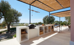 Outdoor kitchen area featuring a modern design with a stone grill and open storage spaces, set under a shaded pergola. Lush greenery and a scenic view enhance the inviting atmosphere, ideal for alfresco dining and entertaining.