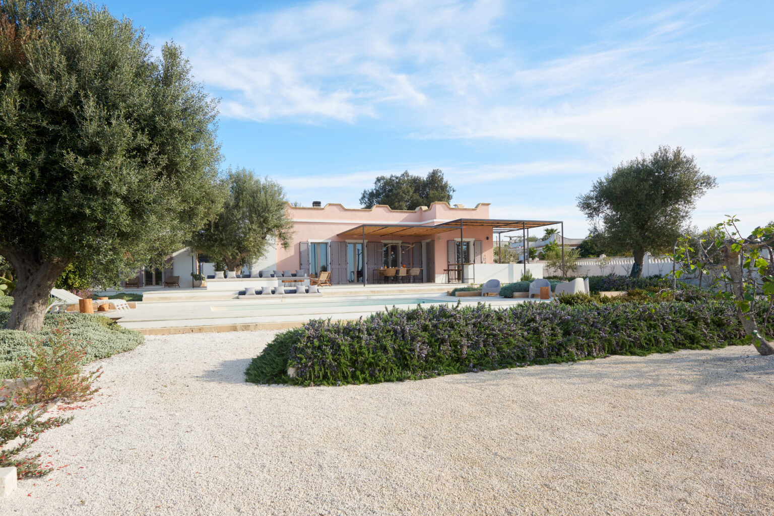 Modern single-story house surrounded by lush greenery and gravel pathways, featuring a spacious outdoor area with sun loungers and a serene landscape under a clear blue sky. | Only on SopranoVillas