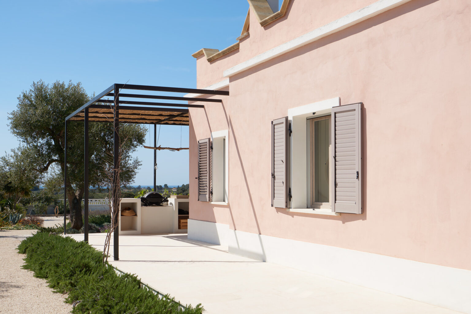 Modern pink house exterior with open shutters, featuring a shaded patio area and outdoor cooking space, surrounded by greenery and clear blue skies. | Only on SopranoVillas