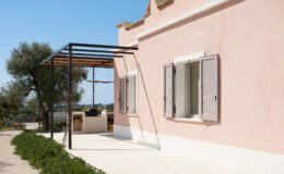 Modern pink house exterior with open shutters, featuring a shaded patio area and outdoor cooking space, surrounded by greenery and clear blue skies.