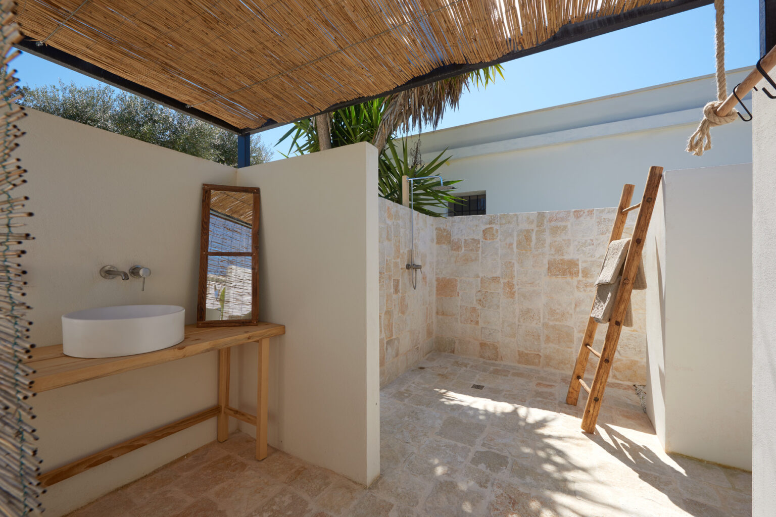 Outdoor shower area featuring natural stone walls, a wooden vanity with a round sink, and a ladder for towels, surrounded by tropical plants and a thatched roof for shade. | Only on SopranoVillas