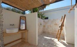 Outdoor shower area featuring natural stone walls, a wooden vanity with a round sink, and a ladder for towels, surrounded by tropical plants and a thatched roof for shade.