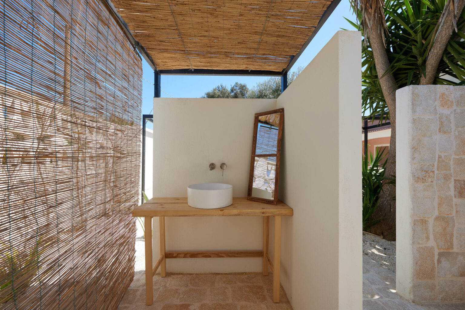 Outdoor bathroom setup featuring a wooden vanity with a round white basin and a mirror, surrounded by bamboo screening and natural greenery, creating a serene and private space. | Only on SopranoVillas
