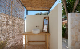 Outdoor bathroom setup featuring a wooden vanity with a round white basin and a mirror, surrounded by bamboo screening and natural greenery, creating a serene and private space.