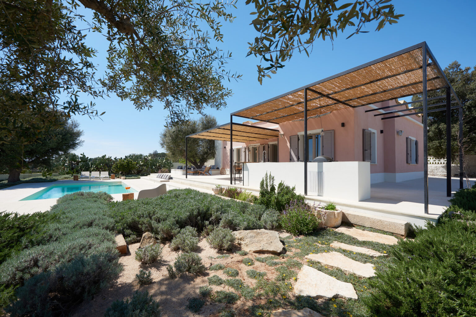 Modern villa with a swimming pool surrounded by lush greenery and stone pathways, featuring a shaded patio area and stylish outdoor seating. Bright blue sky complements the tranquil setting, ideal for relaxation and outdoor living. | Only on SopranoVillas