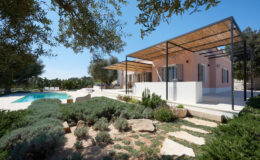 Modern villa with a swimming pool surrounded by lush greenery and stone pathways, featuring a shaded patio area and stylish outdoor seating. Bright blue sky complements the tranquil setting, ideal for relaxation and outdoor living.