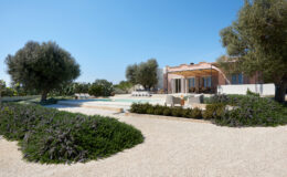 Modern villa with a spacious outdoor area featuring a swimming pool, lounge chairs, and lush landscaping, including olive trees and flowering shrubs, under a clear blue sky.