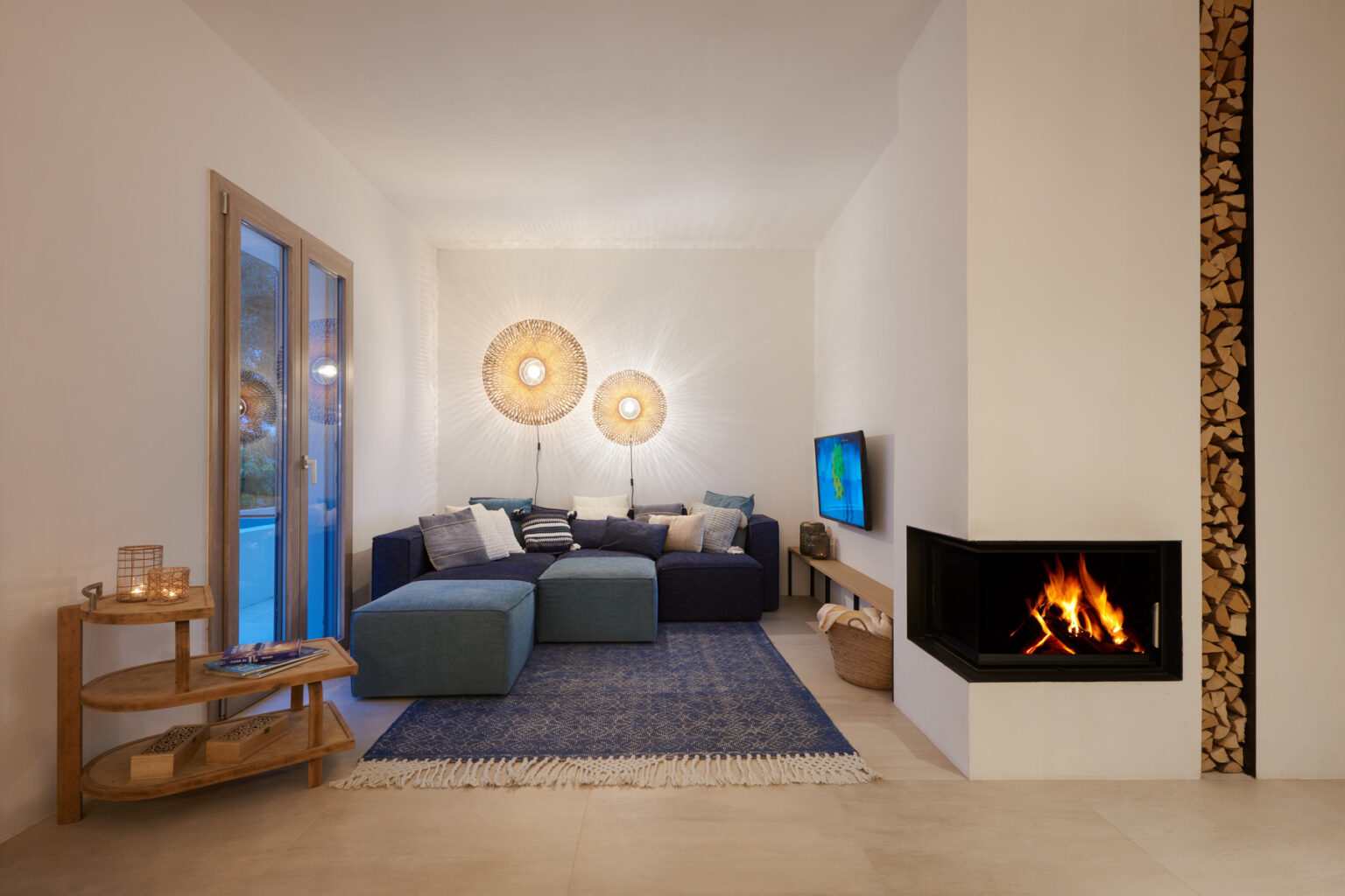 Modern living room featuring a cozy blue sectional sofa, stylish light fixtures, and a contemporary fireplace with stacked wood. The space is decorated with a blue area rug, a wooden side table, and a flat-screen TV, creating a warm and inviting atmosphere. | Only on SopranoVillas