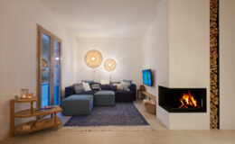 Modern living room featuring a cozy blue sectional sofa, stylish light fixtures, and a contemporary fireplace with stacked wood. The space is decorated with a blue area rug, a wooden side table, and a flat-screen TV, creating a warm and inviting atmosphere.