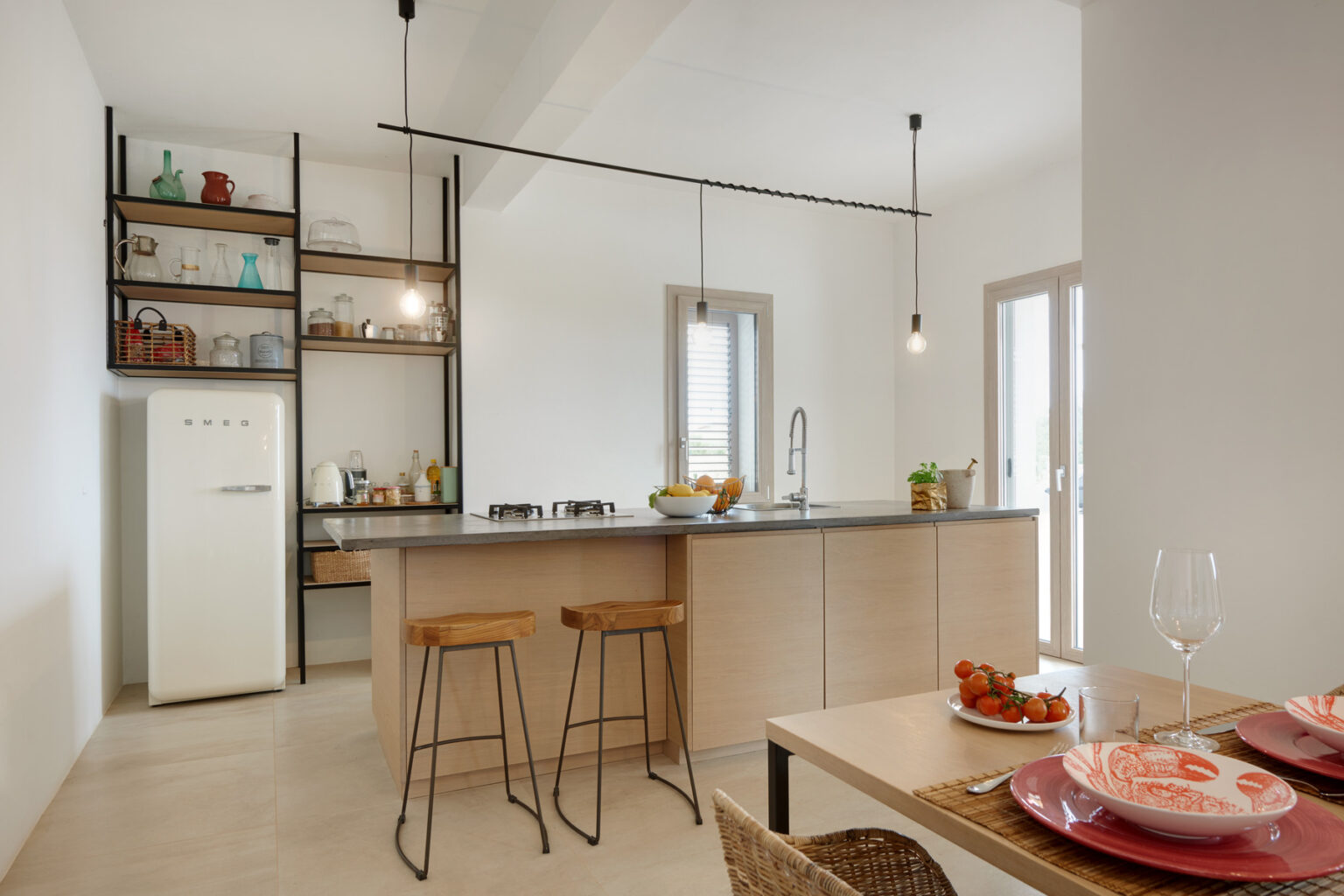 Modern kitchen interior featuring a sleek design with a white SMEG refrigerator, wooden bar stools, and a stylish countertop. The open shelving displays various kitchenware, while natural light streams in through large windows, enhancing the inviting atmosphere. A dining table is set with decorative plates and fresh tomatoes, creating a warm and functional space. | Only on SopranoVillas